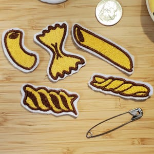 Puede incluir: Parches de pasta bordados en varias formas, incluyendo macarrones con codo, farfalle, penne y rotini, con hilo marrón y amarillo. Un imperdible plateado y una moneda también están en la imagen. Los parches tienen un borde blanco.