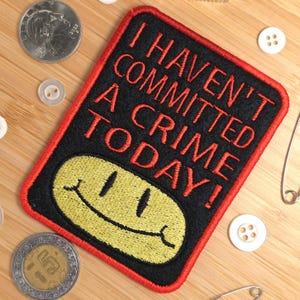 May include: A rectangular embroidered patch with a black background and a red border. The text reads I HAVEN'T COMMITTED A CRIME TODAY! in red capital letters above a yellow embroidered smiley face with a black stitched mouth and eyes.
