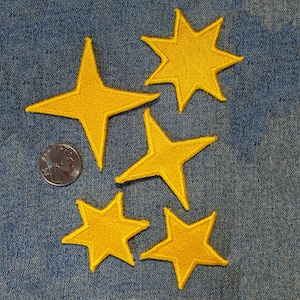 May include: Five yellow embroidered star patches of varying shapes and sizes, including four-pointed and five-pointed designs, arranged alongside a silver-coloured coin for scale, featuring stitched edges and a textured fabric surface.