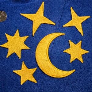 Puede incluir: Un fondo de fieltro azul profundo con parches bordados en amarillo de una luna creciente y cinco estrellas de diversas formas. Una moneda plateada es visible en la esquina superior izquierda, proporcionando una escala para los diseños.