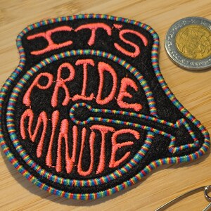 May include: A black embroidered patch with a rainbow-colored border featuring the pink text IT'S PRIDE MINUTE arranged in a circular design with an arrow pointing to the right. A silver coin is positioned to the top right of the patch.