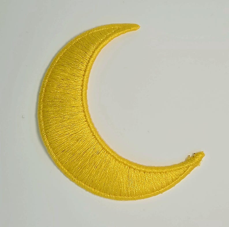 May include: A crescent moon patch featuring bright yellow embroidery thread with a dense, satin-stitch texture. The shape is curved with pointed ends, showcasing a consistent, smooth thread pattern throughout the entire surface of the moon design.