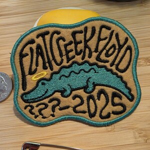 Puede incluir: Parche bordado con el texto "FAT GEEK FLOYD" y un caimán turquesa con un halo. El parche tiene un fondo marrón con un borde turquesa y los números "???-2025".