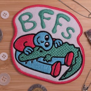 Könnte beinhalten: Ein weißer gestickter Aufnäher mit rosa Rand, der eine blaue Zeichentrickfigur zeigt, die einen grünen Alligator umarmt. Der Text BFFS ist in Grün über den Figuren gestickt. Der Aufnäher ist von weißen Knöpfen, einer Münze und Sicherheitsnadeln umgeben.