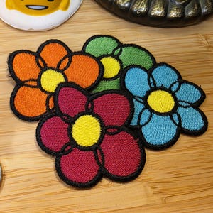 May include: Embroidered flower patches in orange, green, blue, and burgundy, each with a yellow center and black outline. These patches are likely for clothing or craft projects.