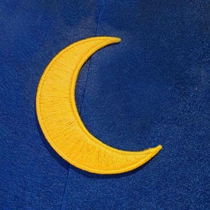 Puede incluir: Un parche de media luna de color amarillo brillante con costuras de bordado densas y uniformes. La forma de la luna está colocada sobre un fondo de tela azul profundo y sólido, creando un contraste visual de un cuerpo celeste en el cielo.