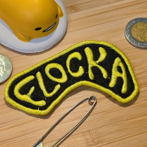May include: A curved, black fabric patch with bright yellow embroidered edges and the yellow embroidered text "FLOCKA" in the center. The patch is accompanied by a silver metal safety pin, a yellow character figurine, and two silver coins.