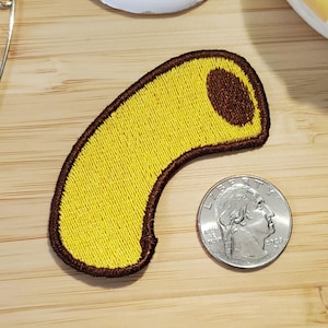 May include: Embroidered patch in the shape of a yellow macaroni noodle with a brown border and a brown hole. A US quarter coin is next to the patch for size comparison.