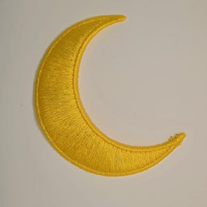 May include: A crescent moon patch featuring vibrant yellow embroidery thread with a dense, satin-stitch texture. The design is shaped as a slender, curved moon with pointed tips, showcasing a consistent, glossy finish across the entire surface.