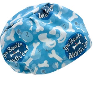 May include: A light blue scrub cap featuring a repeating pattern of white bones and dark blue circular speech bubbles containing the text "You break 'em, we'll fix 'em!" with a small bone graphic and a tiny red heart icon in the center.