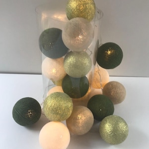 Op de afbeelding: Een lichtsnoer met geweven draadballen in tinten olijfgroen, bosgroen, crème en goud. De verlichte bollen zijn zowel in een doorzichtige cilindervormige houder geplaatst als verspreid rondom de basis van de houder.