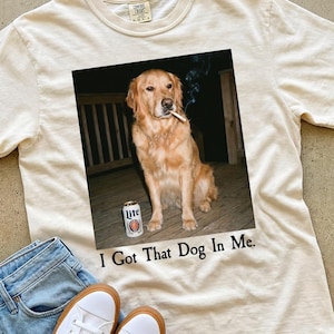 Puede incluir: Camiseta color crema con un gr&aacute;fico de un golden retriever con un cigarrillo y una lata de cerveza. El texto "I Got That Dog In Me" est&aacute; impreso debajo. Vaqueros azul claro y zapatillas blancas completan el look casual.