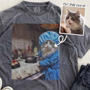 Peut inclure: T-shirt gris foncé avec un graphisme humoristique d'un chat en peignoir bleu et toque de chef, fumant une cigarette tout en cuisinant. Une petite photo d'un chat est incluse. Des baskets grises et un jean noir sont également visibles.