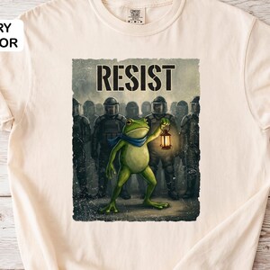 Op de afbeelding: Ivoor kleurig t-shirt met een afbeelding van een groene kikker met een lantaarn, met het woord "RESIST" erboven. De kikker staat voor een groep figuren in zwarte uniformen. Het shirt is gemaakt van zacht materiaal.