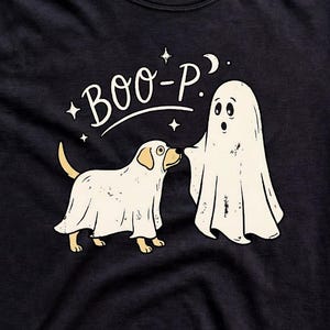 May include: A black short-sleeved t-shirt featuring a graphic of a dog and a ghost wearing white sheets. The text "BOO-P" is printed above the figures, accompanied by small star and moon icons. The shirt has a crew neckline and a standard fit.