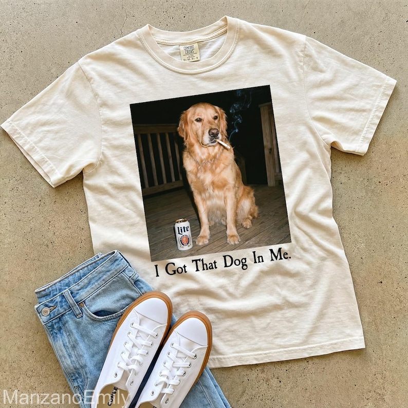 Puede incluir: Camiseta color crema con un gr&aacute;fico de un golden retriever con un cigarrillo y una lata de cerveza. El texto "I Got That Dog In Me" est&aacute; impreso debajo. Vaqueros azul claro y zapatillas blancas completan el look casual.