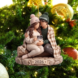 Pode incluir: Um enfeite de Natal com duas figuras sentadas num tronco, uma com um gorro rosa com o nome MIA e a outra com um gorro verde com o nome BENJAMIN. Estão envoltos numa manta xadrez bege e seguram uma ecografia. A base tem a inscrição 2026.