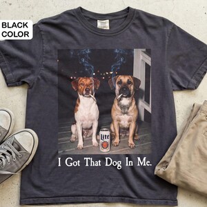 May include: Dark grey t-shirt with a graphic of two dogs smoking cigarettes, with smoke, and a beer can. The text "I Got That Dog In Me" is printed below. The shirt is shown with beige trousers and grey trainers.