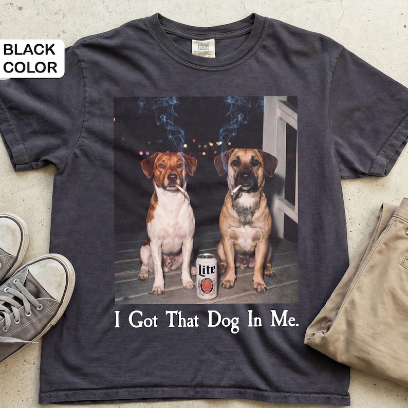 May include: Dark grey t-shirt with a graphic of two dogs smoking cigarettes, with smoke, and a beer can. The text "I Got That Dog In Me" is printed below. The shirt is shown with beige trousers and grey trainers.