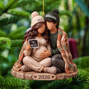 May include: A decorative ornament featuring two figures sitting together on a stone-textured base labeled 2026. One figure wears a beige beanie labeled MIA, the other a dark green beanie labeled BENJAMIN. They are wrapped in a plaid blanket.