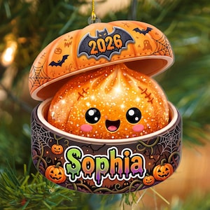 Peut inclure: Une décoration ornementale représentant un personnage de boulette orange pailletée au visage souriant dans un récipient en forme de citrouille. Le couvercle affiche l'année 2026 avec une chauve-souris, et la base porte le nom Sophia en vert.