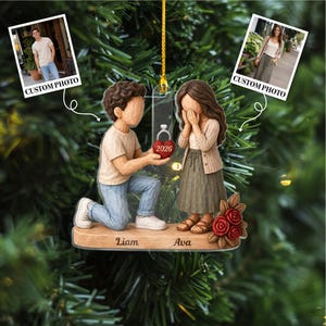May include: A decorative hanging ornament featuring a figurine of a person kneeling to propose to another person who is covering their face with their hands. The base is engraved with the names Liam and Ava, and a red heart displays the year 2026.