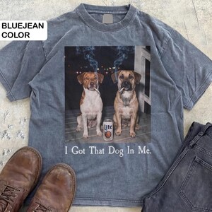 May include: Bluejean-coloured t-shirt with a graphic of two dogs smoking cigarettes, and a can of Lite beer. The text "I Got That Dog In Me" is printed below. The shirt is styled with brown boots and dark jeans.