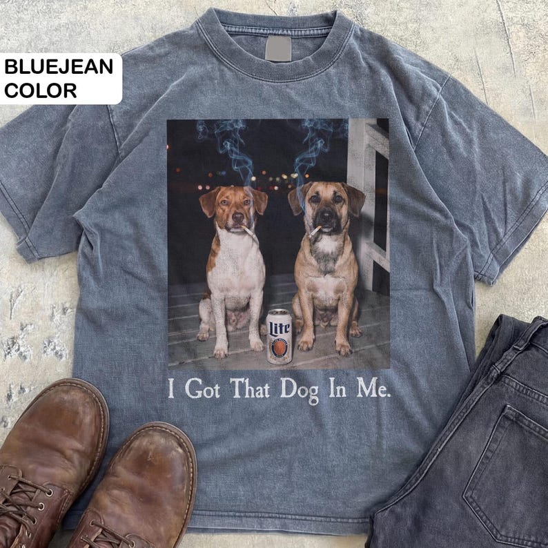 May include: Bluejean-coloured t-shirt with a graphic of two dogs smoking cigarettes, and a can of Lite beer. The text "I Got That Dog In Me" is printed below. The shirt is styled with brown boots and dark jeans.
