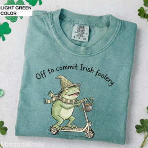 May include: Light green t-shirt featuring the phrase "Off to commit Irish foolery" and a cartoon frog on a scooter. The frog is wearing a hat and scarf, and holding a pot of gold and shamrocks. The shirt has a crew neck.