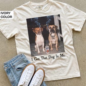May include: An ivory-coloured t-shirt with a graphic of two dogs smoking cigarettes and a beer can. The text "I Got That Dog In Me" is printed below. The shirt is shown with blue jeans and white trainers.