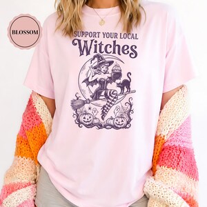 May include: A light pink t-shirt featuring a dark purple graphic print with the text SUPPORT YOUR LOCAL WITCHES. The design depicts a figure in a witch hat riding a broomstick with a crescent moon, a black cat, pumpkins, and ghosts.