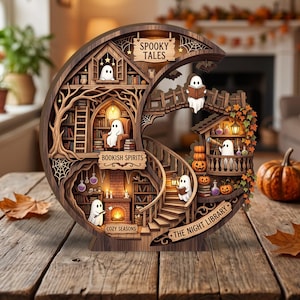 May include: A crescent-shaped wooden layered display featuring a library scene with ghosts, bookshelves, a fireplace, and pumpkins. Text includes "SPOOKY TALES," "BOOKISH SPIRITS," "COZY SEASONS," and "THE NIGHT LIBRARY" engraved on the wooden structure.