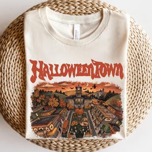 May include: A cream-colored crew neck t-shirt featuring the red text HALLOWEENTOWN above a detailed illustration of a town square at dusk, including a city hall building, a large pumpkin, autumn trees, flying bats, and a witch on a broomstick.