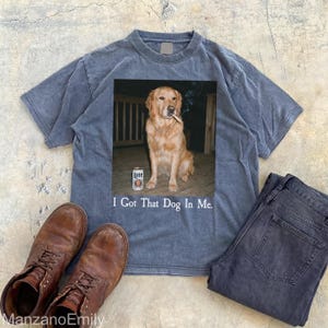 Puede incluir: Camiseta azul con un gr&aacute;fico de un golden retriever con un cigarrillo y una lata de cerveza. El texto "I Got That Dog In Me" est&aacute; debajo. Botas marrones y vaqueros oscuros tambi&eacute;n est&aacute;n en la imagen.