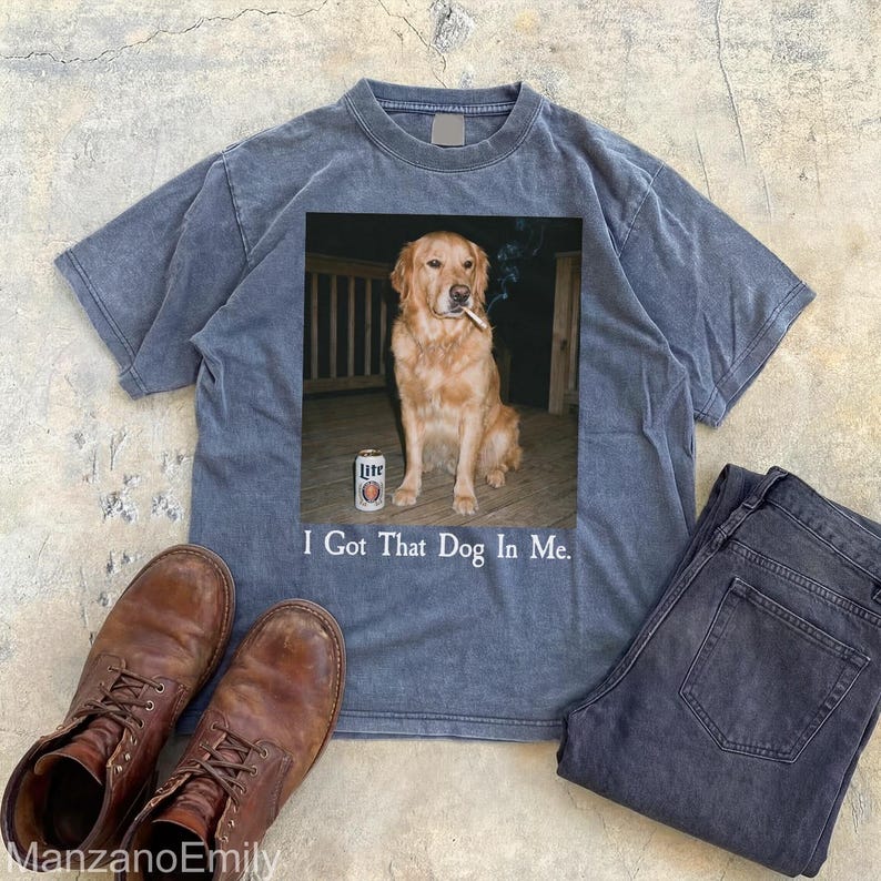 Puede incluir: Camiseta azul con un gr&aacute;fico de un golden retriever con un cigarrillo y una lata de cerveza. El texto "I Got That Dog In Me" est&aacute; debajo. Botas marrones y vaqueros oscuros tambi&eacute;n est&aacute;n en la imagen.