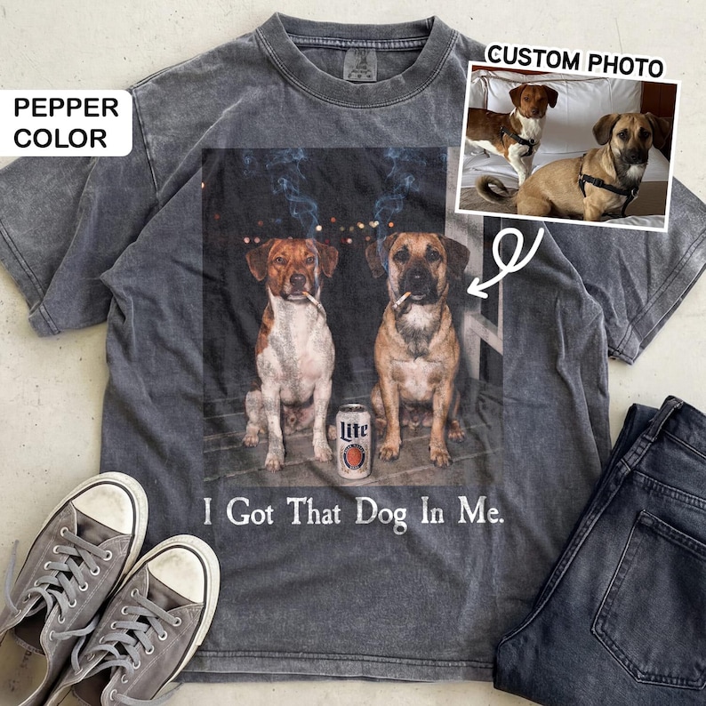 May include: Pepper-coloured t-shirt with a graphic of two dogs smoking cigarettes, and a beer can. The text "I Got That Dog In Me" is printed below the image. Grey trainers and jeans are next to the shirt.