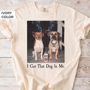 May include: Ivory t-shirt with a graphic of two dogs smoking cigarettes, a beer can, and the text "I Got That Dog In Me." The dogs are brown and white, and brown. The beer can is white and orange.