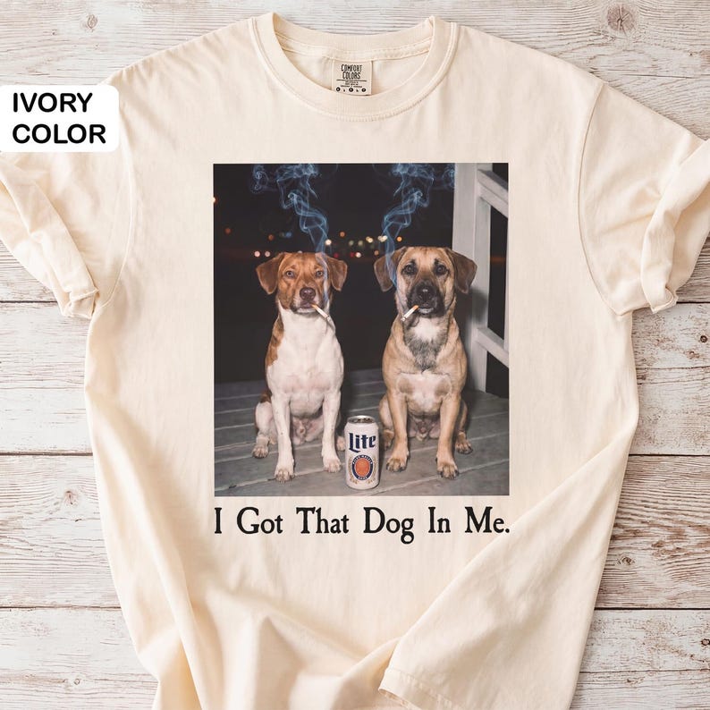 May include: Ivory t-shirt with a graphic of two dogs smoking cigarettes, a beer can, and the text "I Got That Dog In Me." The dogs are brown and white, and brown. The beer can is white and orange.