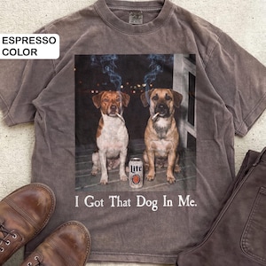 May include: Espresso-coloured t-shirt with a graphic of two dogs smoking cigarettes and a can of Lite beer. The text "I Got That Dog In Me" is printed below. The shirt is shown with brown trousers and shoes.