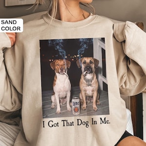 May include: Sand-coloured sweatshirt with a graphic of two dogs smoking cigarettes, with smoke rising above their heads. A can of Lite beer sits between them. The text "I Got That Dog In Me" is printed below the image.