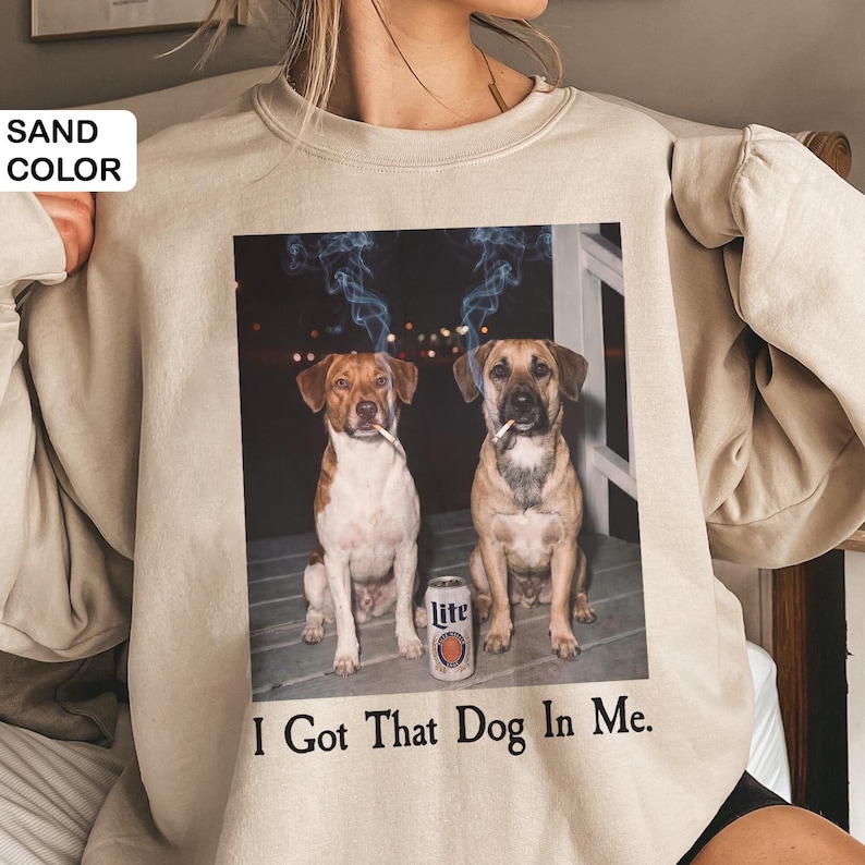 May include: Sand-coloured sweatshirt with a graphic of two dogs smoking cigarettes, with smoke rising above their heads. A can of Lite beer sits between them. The text "I Got That Dog In Me" is printed below the image.