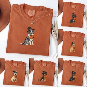 May include: A rust-colored crew neck t-shirt featuring a graphic of a dog wearing a witch hat and holding a jack-o'-lantern. The shirt has a Comfort Colors label at the inner neckline and is displayed alongside various dog breed variations.