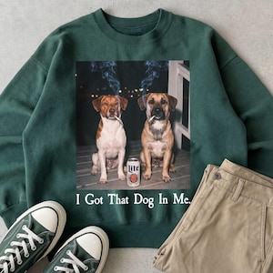 May include: Dark green sweatshirt featuring a graphic of two dogs smoking cigarettes, with a can of Lite beer between them. The text "I Got That Dog In Me" is printed below the graphic. Green trainers and khaki trousers are also visible.
