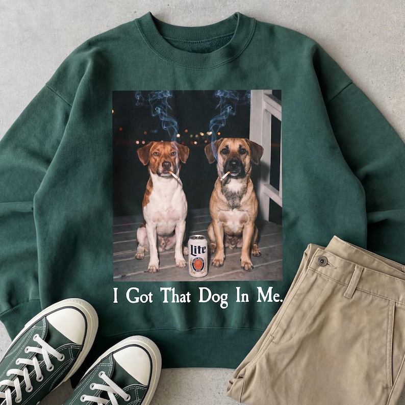 May include: Dark green sweatshirt featuring a graphic of two dogs smoking cigarettes, with a can of Lite beer between them. The text "I Got That Dog In Me" is printed below the graphic. Green trainers and khaki trousers are also visible.