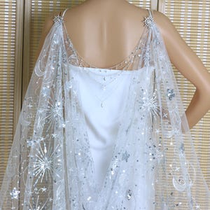 May include: A sheer, white cape with celestial embellishments, including stars and crescent moons, is draped over a white dress. The cape is secured with star-shaped brooches at the shoulders and adorned with silver sequins.
