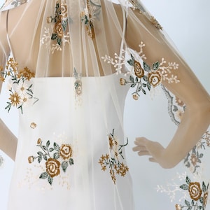 May include: An ivory wedding veil with floral embroidery in gold, green, and white. The veil is draped over a white dress, with the floral design accenting the edges and cascading down the fabric. The overall effect is delicate and romantic.