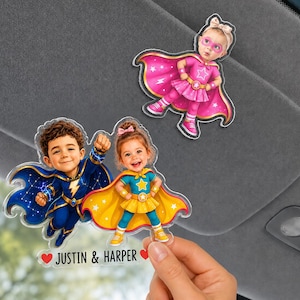 Pode incluir: Pingentes de carro em acrílico com tema de super-heróis, apresentando ilustrações de crianças em trajes de super-heróis. Os pingentes são em tons de azul, amarelo e rosa, com os nomes "JUSTIN & HARPER" impressos em um deles. Os pingentes são segurados por uma mão.