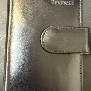 May include: Black leather wallet with a flap closure. The wallet features the embossed letters "GB" above the word "GOLDBACK". The wallet has a stitched border and a smooth, glossy finish.