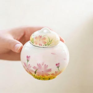 May include: A small, round, white ceramic jar with a lid, hand-painted with pink and yellow floral designs. The jar has a small loop on top of the lid. The jar is held in a person's hand.