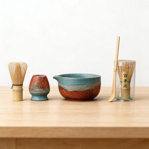 May include: A matcha tea set featuring a bamboo whisk, a ceramic bowl with a blue and brown glaze, a small ceramic holder, a bamboo scoop, and a container with a bamboo whisk. The set is arranged on a wooden surface.